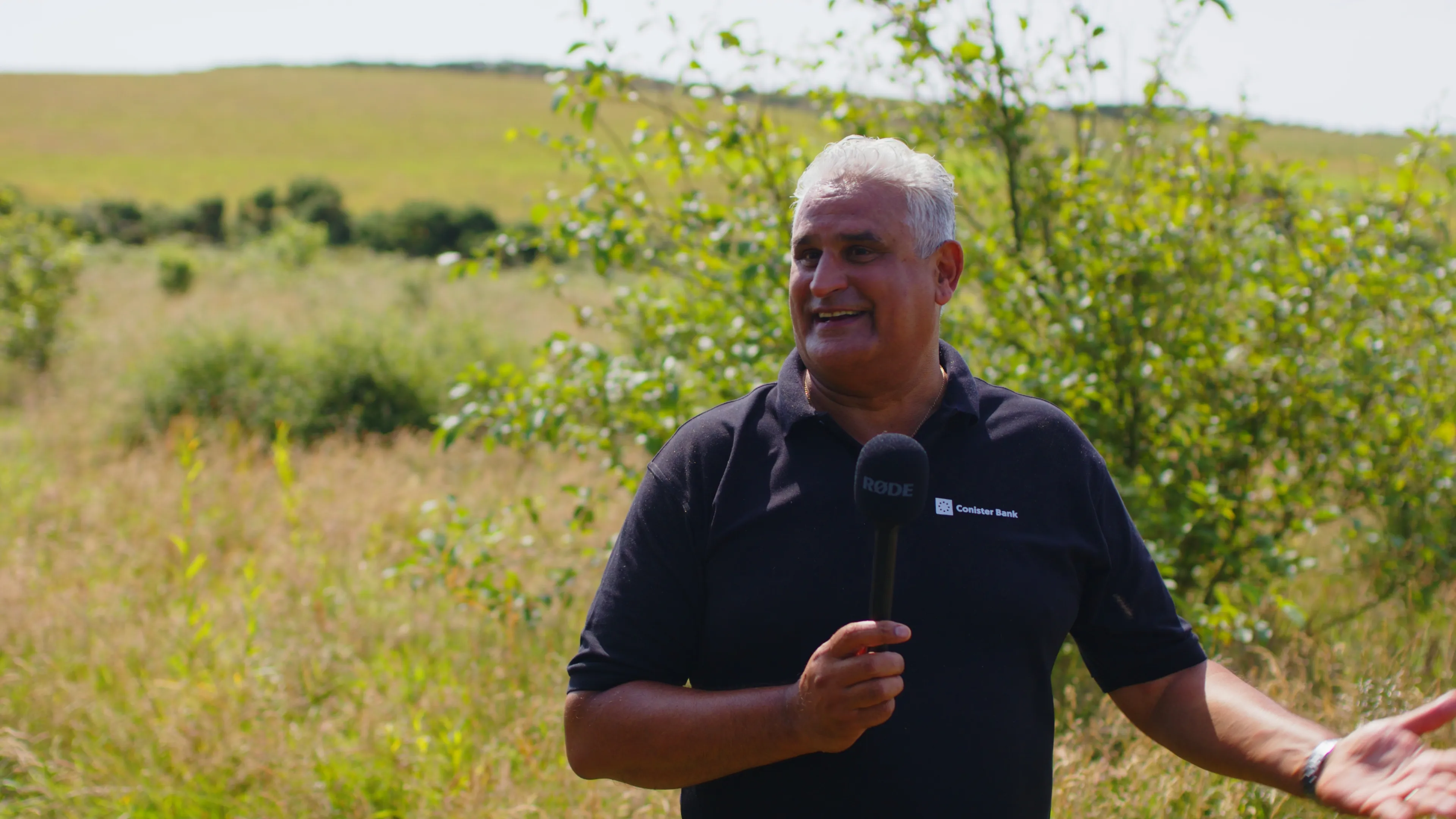 A man stands outdoors in a grassy field, smiling and holding a microphone. The sunny landscape features lush greenery and gentle hills.
